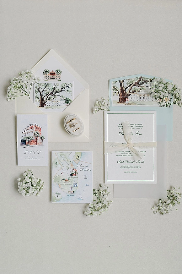 Invitation suite with illustrated cards, envelopes, ribbon tie, wedding rings, and baby’s breath arranged on a light gray surface