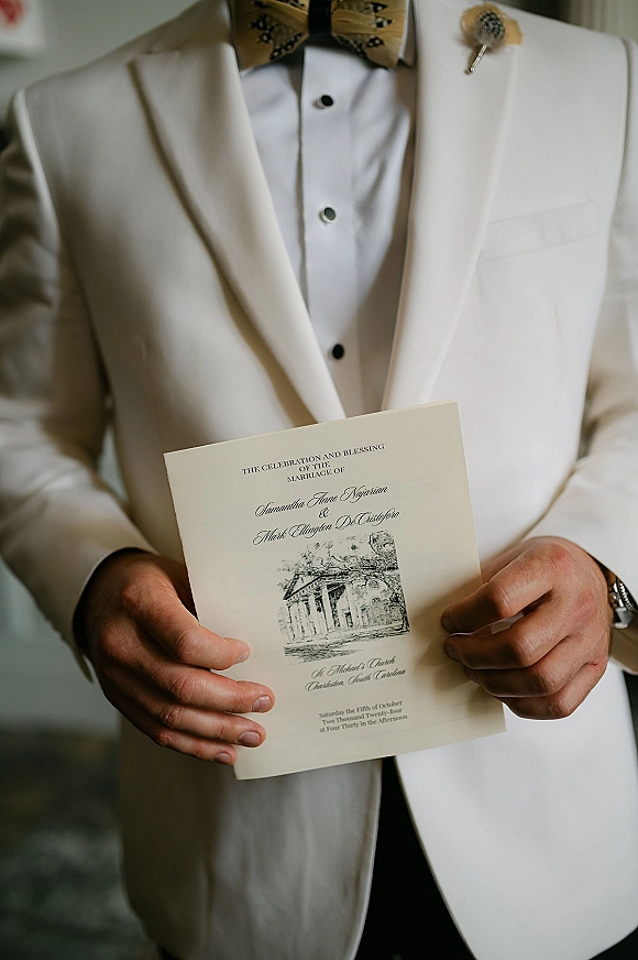 Wedding invitation held by groom in a white tuxedo jacket with bow tie, boutonniere, and wristwatch against a neutral indoor background