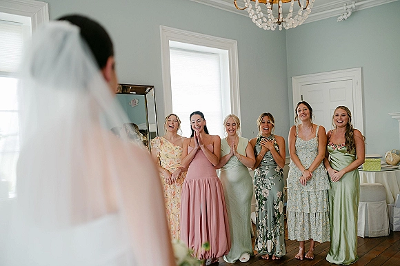 Bridesmaid first look as bridesmaids reaction unfolds when the bride reveals her veil in a bright getting ready room with mirror and chandelier