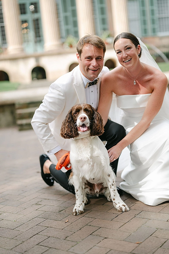 Couple portrait of bride and groom with dog, bride in strapless dress and veil, groom in white tuxedo, in a brick courtyard