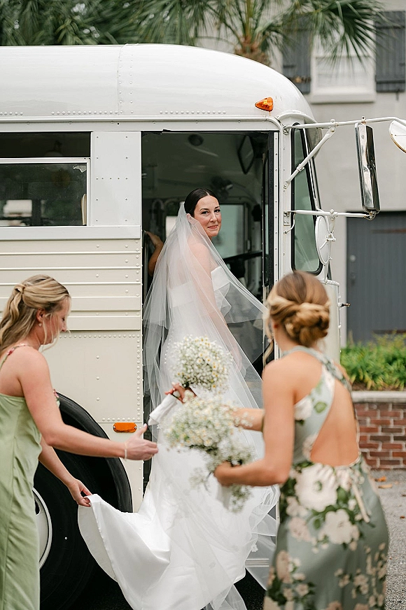 Bride arrival as she steps off a vintage bus, cathedral veil flowing while bridesmaids assist, holding a baby's breath bouquet by palm trees