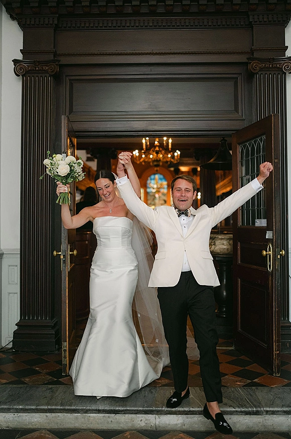 Wedding recessional as bride and groom cheer with raised hands, bouquet and veil flowing as they stride through dark wood doors under a chandelier