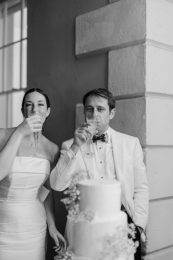Wedding toast as bride and groom sip from champagne flutes beside a two-tier white cake against a stone column and window backdrop