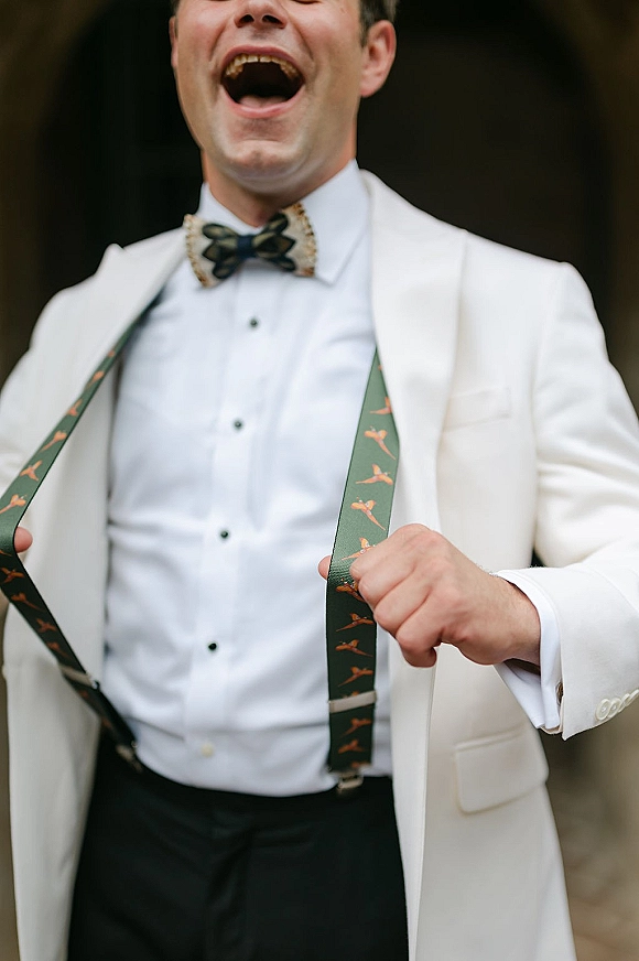 Groom portrait of a laughing groom in a white tuxedo jacket with bow tie and suspenders, standing before softly blurred greenery outdoors