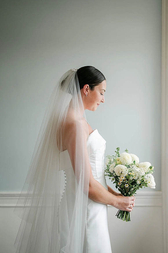 Bridal portrait of a side profile bride in a strapless button-back gown, long veil, holding a white-and-green bouquet against neutral wainscoting