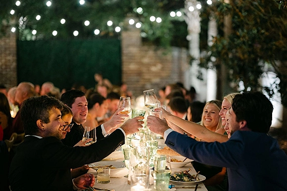 Wedding reception toast as guests raise champagne flutes over a long table with candlelight accents under string lights on an outdoor patio