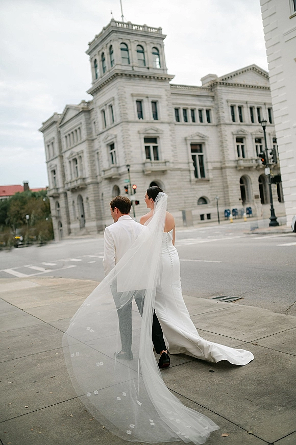 Couple portrait of bride and groom walking, seen from behind as her long veil trails over a city crosswalk by a historic building