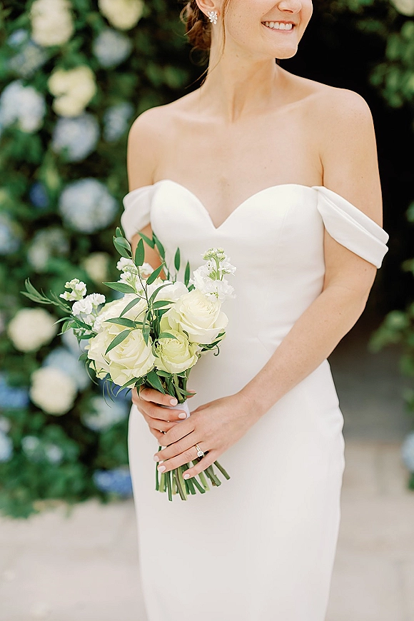 Bridal portrait of a smiling bride in an off the shoulder wedding dress holding white rose and greenery bouquet against a floral wall backdrop