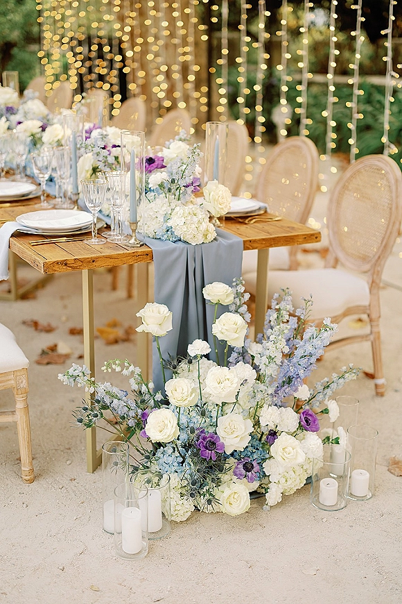 Reception tablescape with blue table runner, gold flatware, taper candles and lush centerpiece on a wood banquet table under string lights in a garden