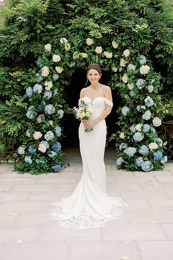 Bridal portrait of a bride in a strapless wedding dress holding a white rose bouquet beneath a hydrangea arch on a garden walkway