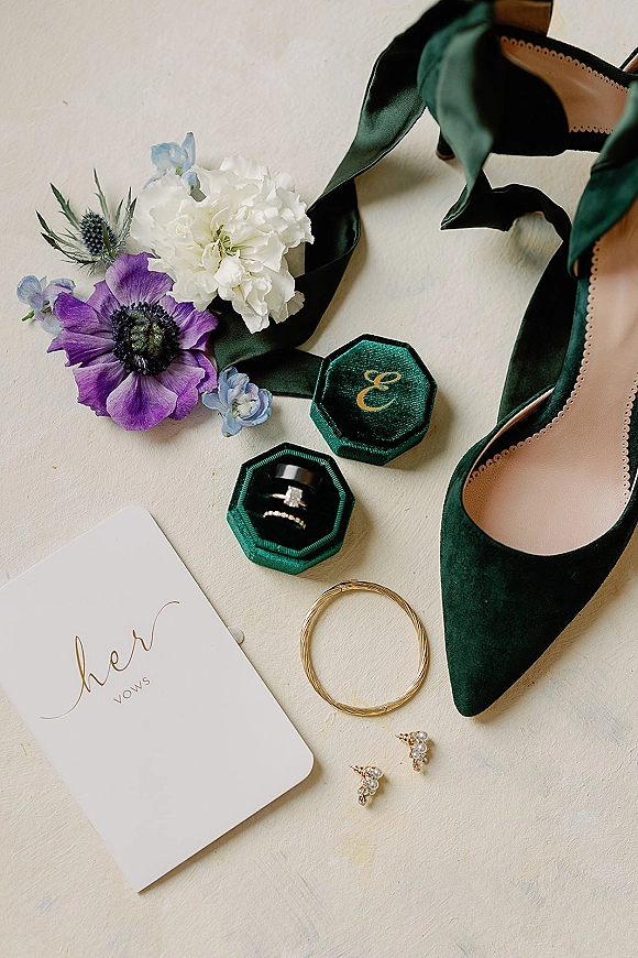Wedding flat lay with wedding detail flat lay styling, green velvet ring box, rings, vow book, pearl earrings, green heels on textured surface
