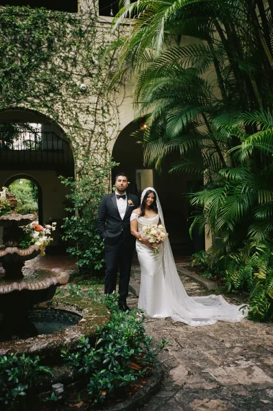 Couple portrait of the bride and groom standing by the fountain, with the bride holding a bouquet amid tropical greenery and courtyard arches.