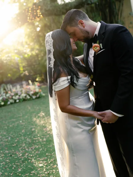 Couple portrait of a bride and groom holding hands, her lace veil catching the light in a sunlit garden under tall trees.