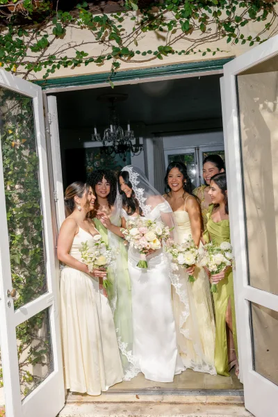 Bride and bridesmaids in sage green bridesmaid dresses stand with bouquets by French doors framed in vines beneath a chandelier.