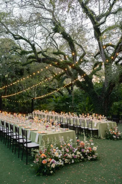Reception tablescape showcasing refined outdoor reception decor, with taper candles glowing along long tables amid garden trees and greenery.