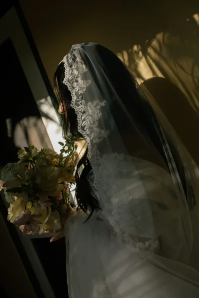 Bridal portrait of a bride with veil, her scalloped lace veil draped over one shoulder as she gazes by a window.