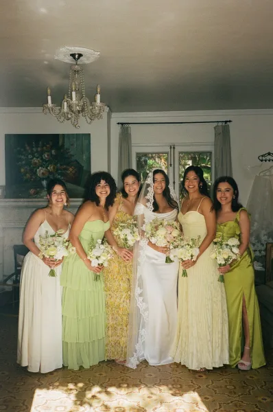 Bride and bridesmaids in mix and match bridesmaid dresses smile together by a tall window in an elegant room with chandelier and fireplace