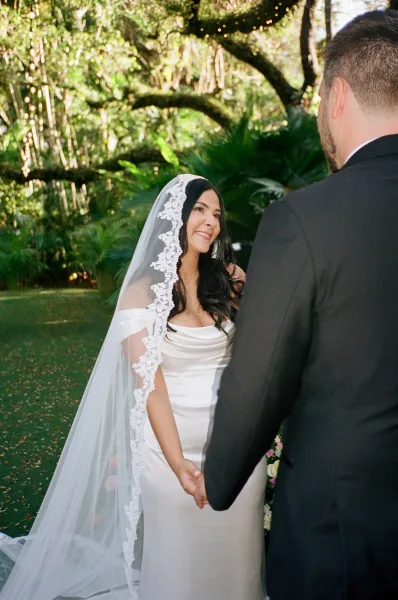 Ceremony moment in a wedding vows exchange as the bride and groom hold hands, her lace veil trailing while they stand beneath garden trees.