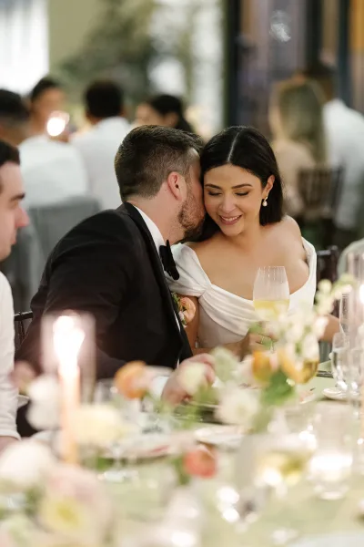 Reception couple moment with the bride and groom whispering at a dinner table, the bride lifting a wine glass amid an indoor reception and greenery.