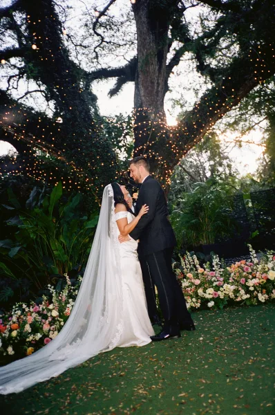 Couple portrait as the bride and groom embrace beneath a large tree, under warm string lights, with garden foliage at dusk on the lawn.