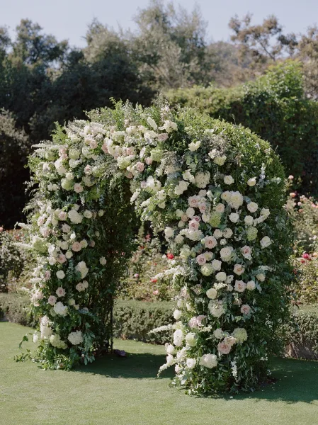 Floral wedding arch with white and blush roses, hydrangeas, and greenery garland on a garden lawn under blue sky