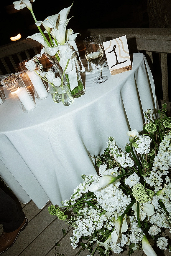 Reception tablescape with white wedding tablescape details, calla lilies in bud vases, cylinder candles, wine glasses and place cards on a wood stand by a stone wall