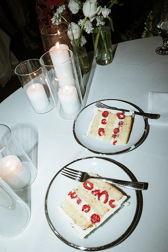 Wedding cake slices with raspberries and white frosting on silver-rim plates with forks, beside pillar candles and tulip bud vases in a dark reception room