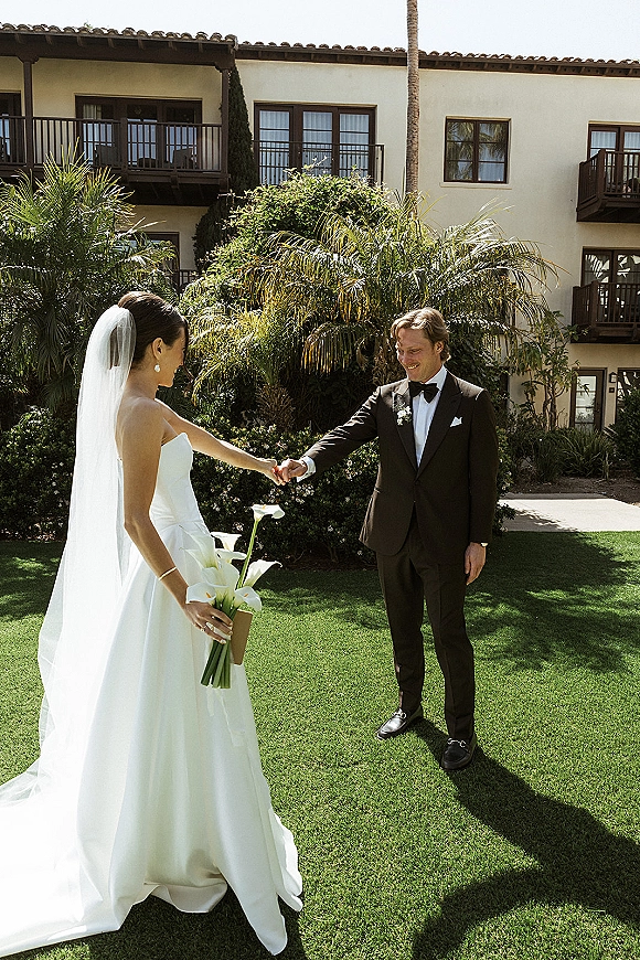 Wedding couple portrait of bride and groom holding hands, her long veil and calla lily bouquet, on a sunny lawn with palm trees