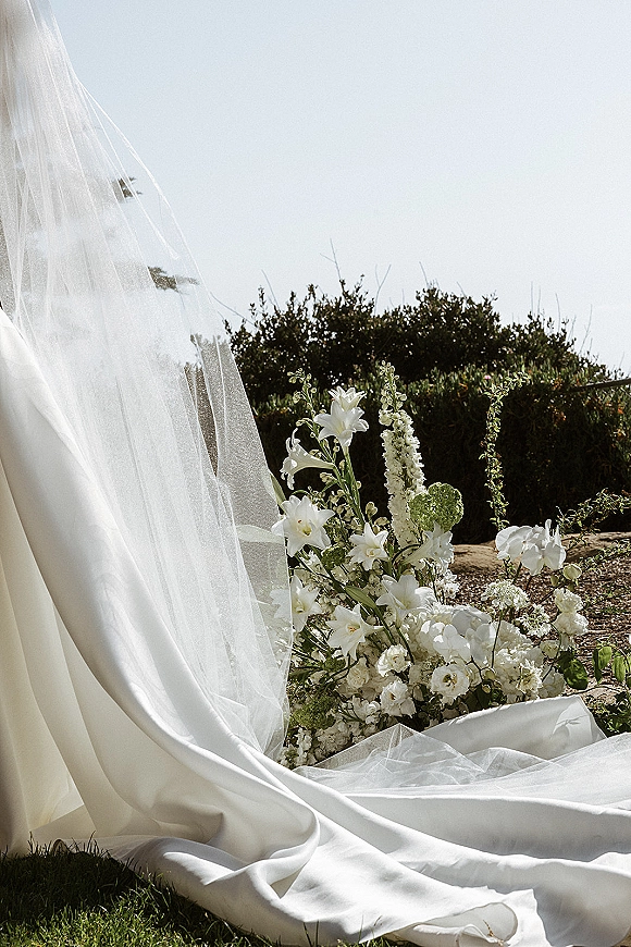 Wedding dress detail with bridal veil detail draped in tulle beside white lilies and greenery along a garden path
