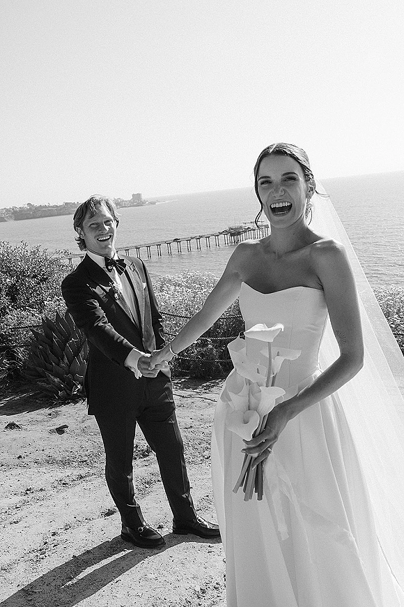Couple portrait in black and white wedding photo, bride and groom holding hands, laughing by the ocean coastline with pier behind