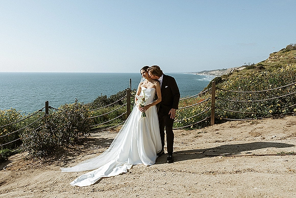Couple portrait of bride and groom outdoors, groom kissing her cheek as she holds calla lilies beside ocean cliffs and rope fence
