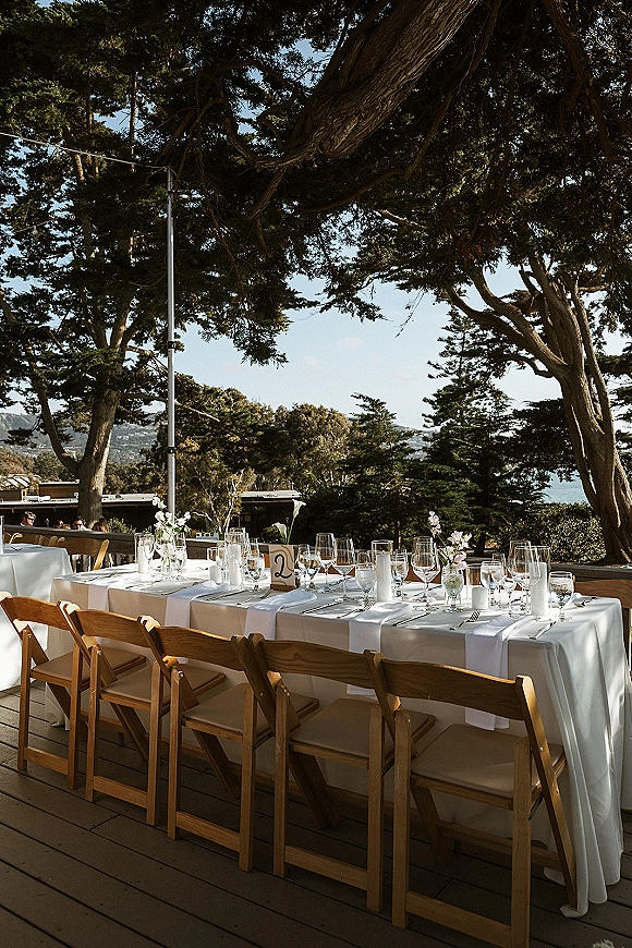 Reception tablescape with white linen runner, wood folding chairs, bud vase flowers and taper candles under string lights on a coastal deck