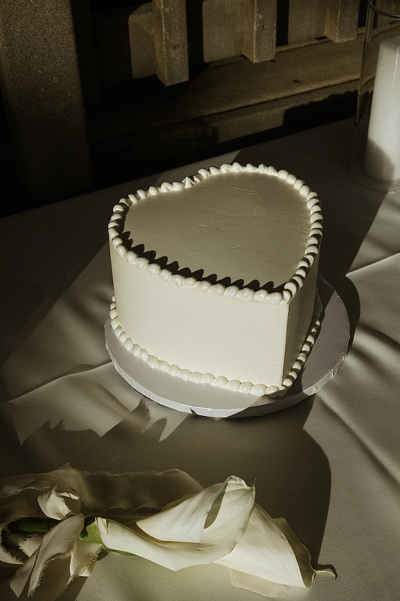 Wedding cake with heart shaped wedding cake design, white buttercream and piped border, beside calla lilies on a tablecloth by a sunlit wood wall