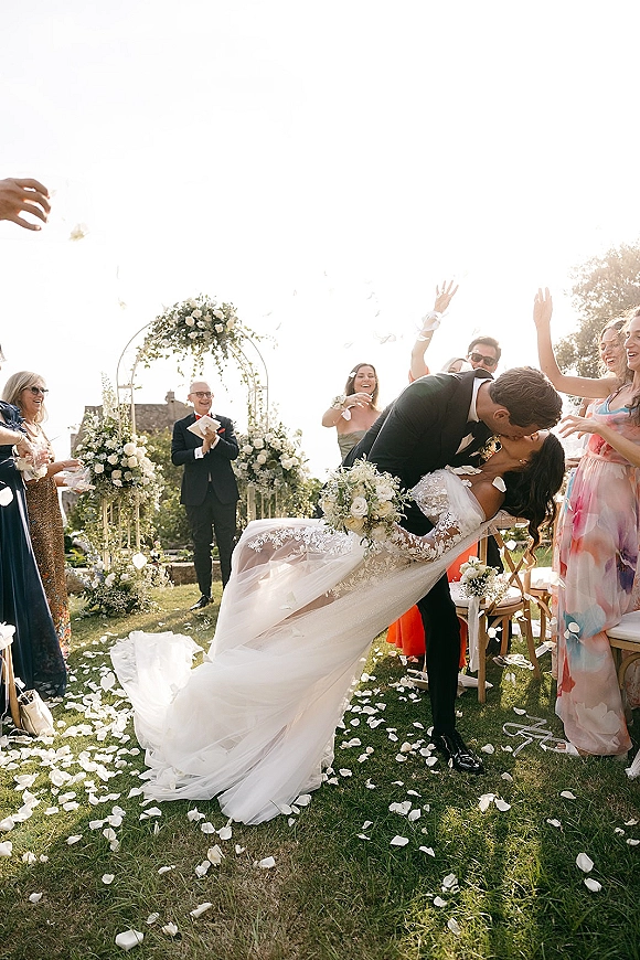 Wedding kiss as the groom dips the bride in a lace dress holding a bouquet, with confetti flying down the garden aisle under a floral arch