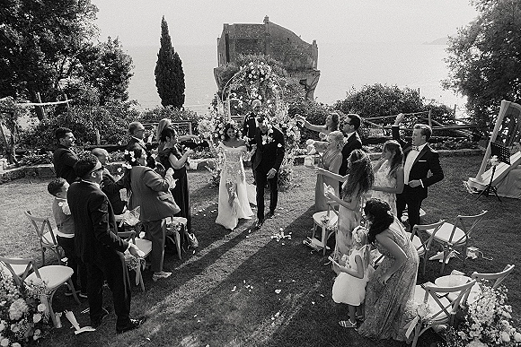 Wedding recessional as bride and groom walk down the aisle through a flower petal toss, with floral arch, guests, and ocean-view lawn backdrop