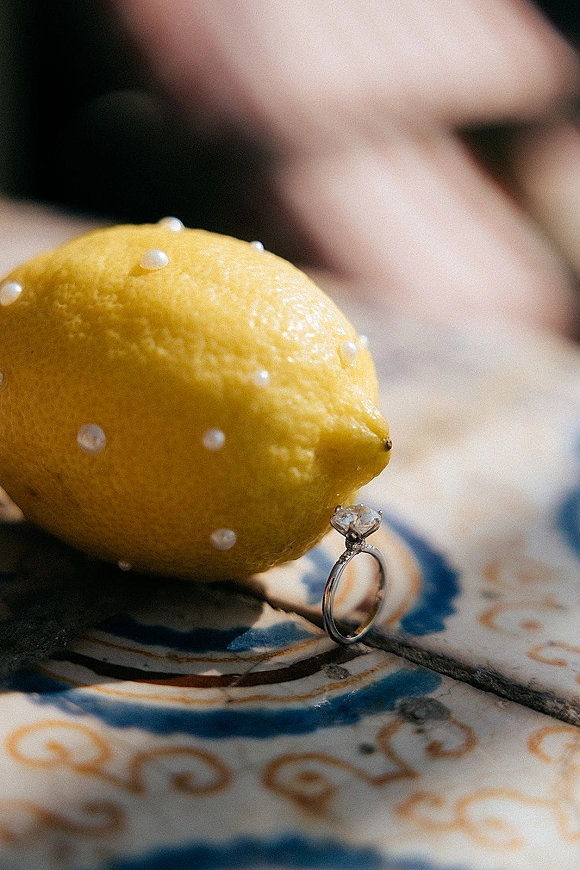 Engagement ring with a diamond solitaire ring beside a lemon and sugar pearls on patterned tile, with blurred hands in the background
