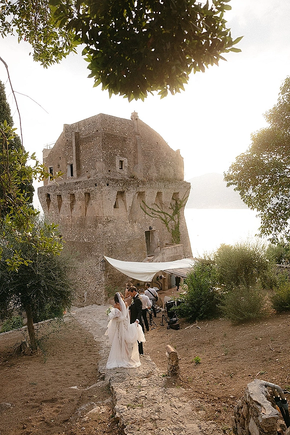 Wedding couple portrait of bride and groom kissing under a white fabric canopy, her veil and bouquet by a stone tower path at golden hour.
