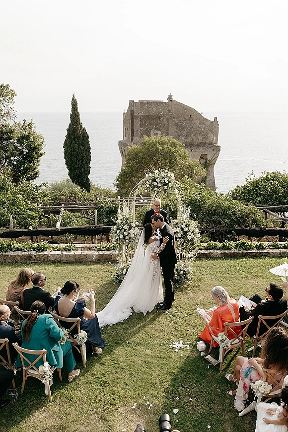 Ceremony kiss at an outdoor wedding ceremony under a white floral arch, bride in long veil and groom in tux with ocean behind