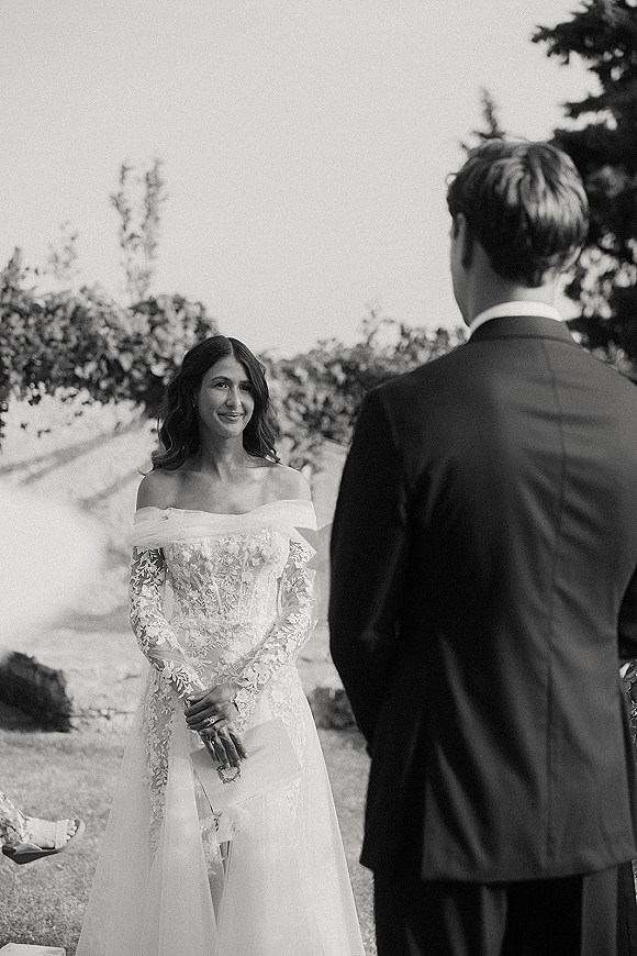 Wedding vows as bride in off-the-shoulder lace dress reads from an envelope to groom in a garden with stone wall backdrop
