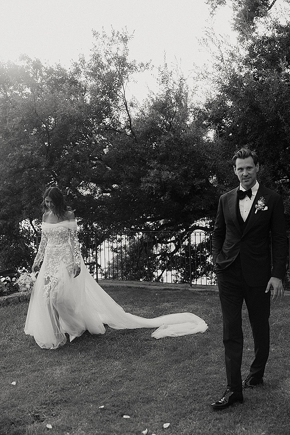 Wedding couple portrait in black and white, bride in off-shoulder lace gown with long train and bouquet beside groom in tux on garden lawn