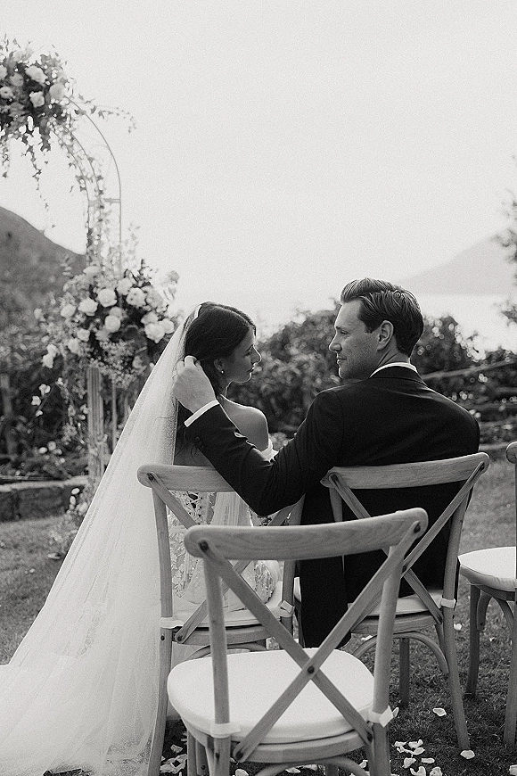 Ceremony moment as the groom adjusts the bride’s wedding veil while they sit under a floral arch in an outdoor garden setting