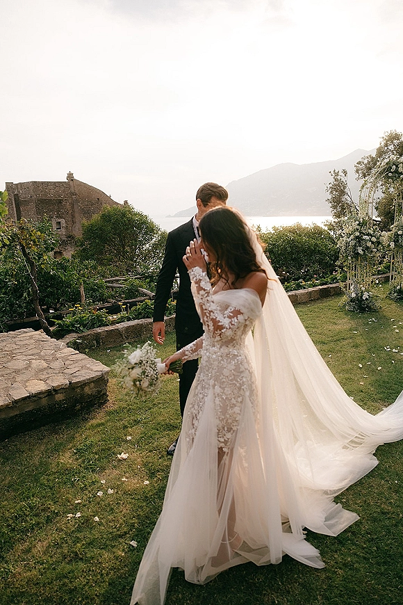 Wedding couple portrait of bride and groom standing on a stone terrace, her long veil trailing, with floral arch and ocean view behind