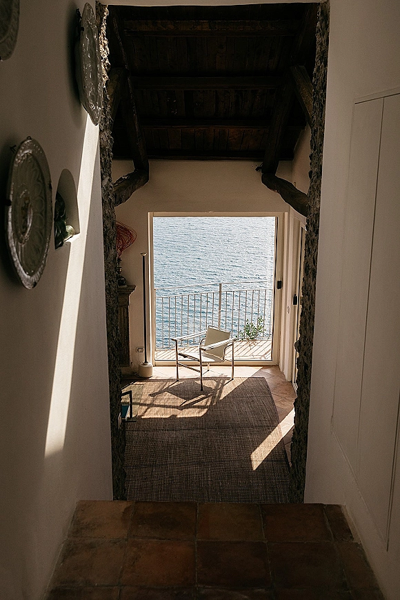 Bridal suite interior with a woven rug and armchair, sunlight casting shadows on white walls, glass doors opening to an ocean-view balcony