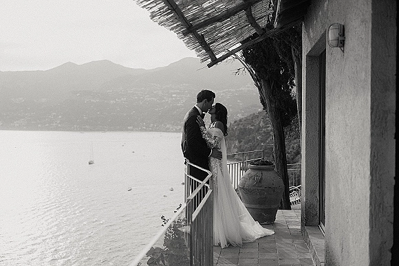 Wedding kiss portrait of bride and groom kiss, her lace sleeves and long veil flowing on a terrace balcony with lake and mountains behind