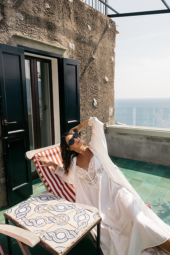 Bridal portrait of a bride in sunglasses lounging on a terrace chair, lace wedding dress under robe, with ocean view behind