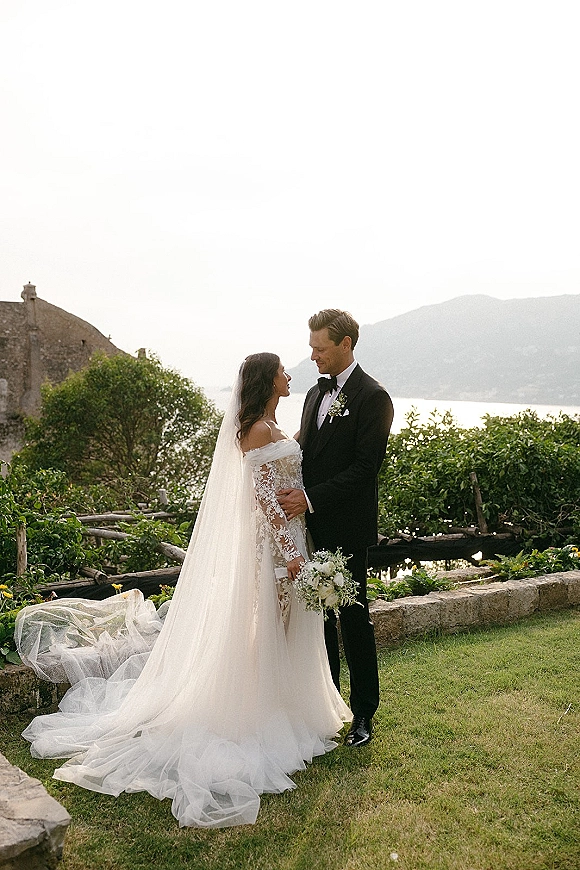 Couple portrait of bride and groom holding hands, her off-shoulder lace gown with long veil and white bouquet in a garden by a stone wall
