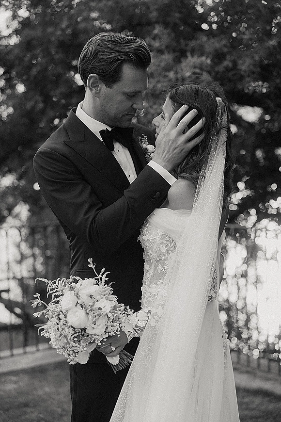 Wedding couple portrait in black and white as bride and groom embrace, his hand on her face, bouquet and veil in a garden bokeh backdrop