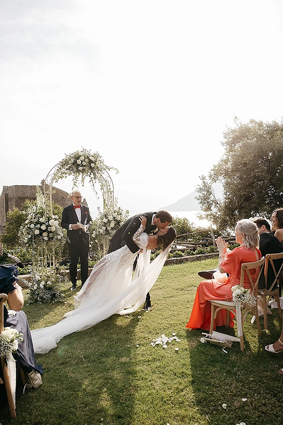 Ceremony kiss under a floral arch as groom dips the bride, veil and long train flowing on a seaside lawn with guests applauding