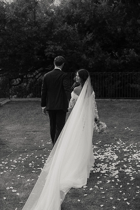 Couple portrait of bride and groom walking away hand in hand, bride looking back as long veil trails over rose petals on grass lawn