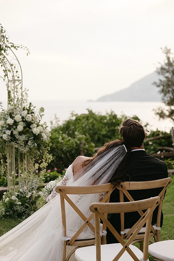 Couple portrait of bride leaning on groom, seated with lace dress and veil train under a white rose arch, ocean and hills behind